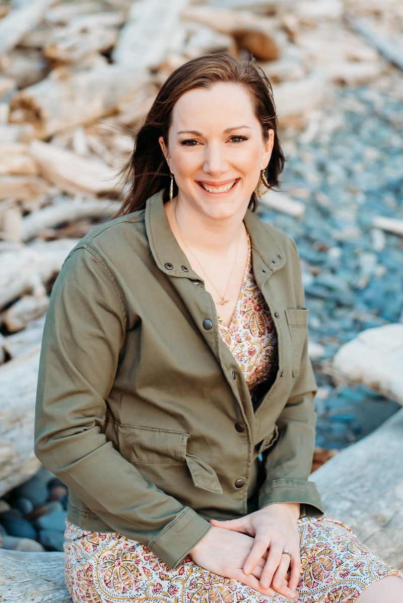 Pearl Stokes smiling, seated on driftwood at a pebble beach